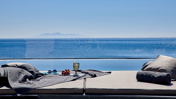 A sun lounger with a view of the sea and mountains, a glass of water, and a plate of grapes. - Incognito Villa (Santorini)