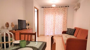 A living room with a sofa, a dining table with a plate and a green mug, a TV on a wooden cabinet, and a floral curtain. - Acv - costa marina I-1 línea planta 5 sur (Oropesa del Mar)