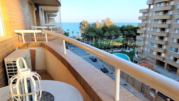 A balcony with a view of a road and buildings, a white outdoor table with a decorative item, and a building with a mesh structure. - Acv - costa marina I-1 línea planta 5 sur (Oropesa del Mar)