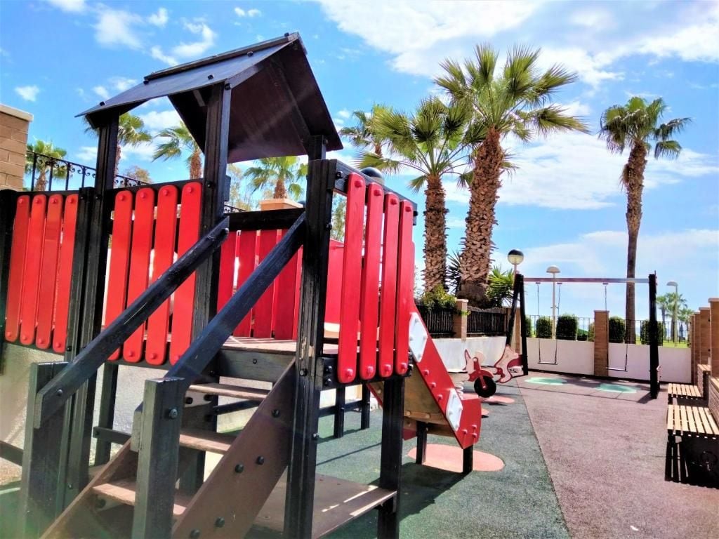 A playground with red and black play structures, including a slide and climbing area, surrounded by palm trees and a clear sky.