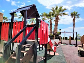 A playground with red and black play structures, including a slide and climbing area, surrounded by palm trees and a clear sky. - Acv - costa marina I-1 línea planta 5 sur (Oropesa del Mar)