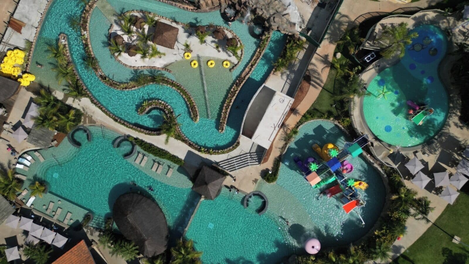 A large outdoor swimming pool area with multiple sections, including a central winding pool and a smaller circular pool, surrounded by lounge areas and palm trees.