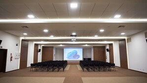 Une salle de conférence avec des rangées de chaises disposées face à un écran de projection, un pupitre et un bureau pour l'orateur.