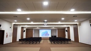 Une salle de conférence avec des rangées de chaises disposées face à un écran de projection, un pupitre et un bureau pour l'orateur.