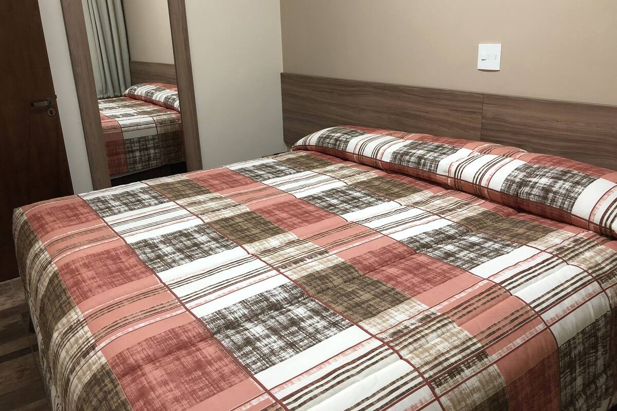 Una cama bien hecha con colcha y almohadas a cuadros, un cabecero de madera, un espejo y un interruptor de luz en la pared.