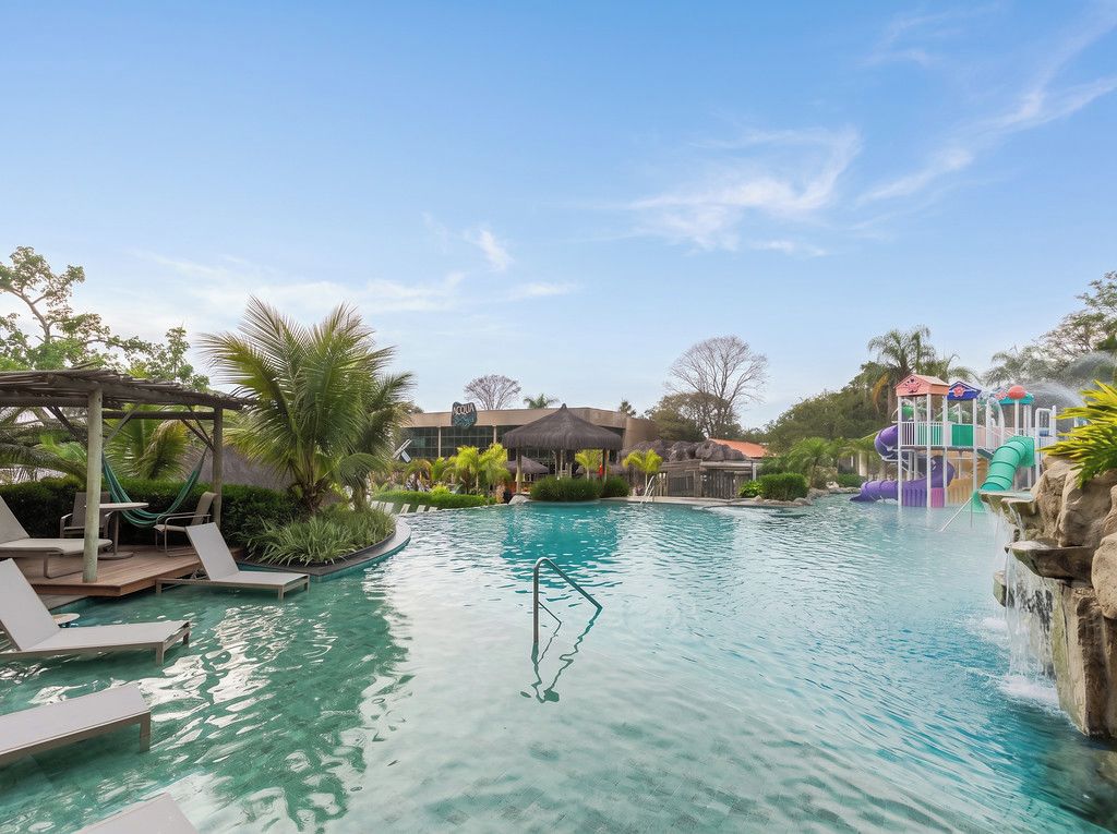 Une piscine avec des chaises longues, un toboggan aquatique et, au fond, un bâtiment dont le toit est recouvert de chaume.