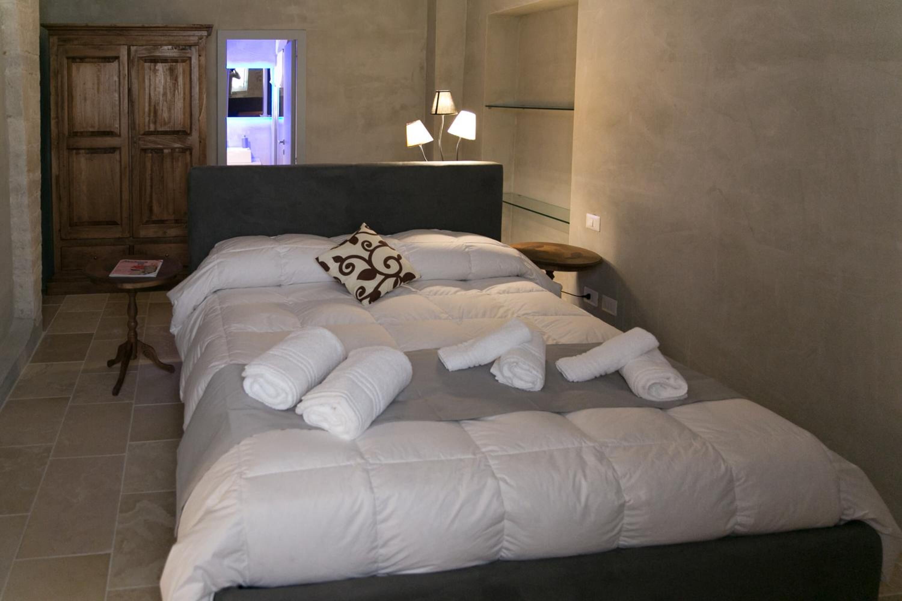 Una cama bien hecha con ropa de cama blanca y toallas enrolladas, una mesita de noche con lámpara, y un armario de madera en una habitación con pared lisa y suelo de baldosas.