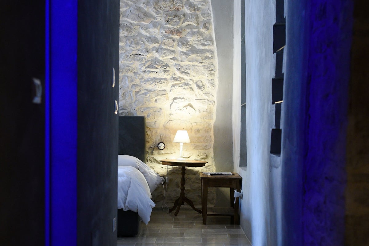 A narrow hallway with a stone wall, a bed with white bedding, a small table with a lamp, and a blue light.