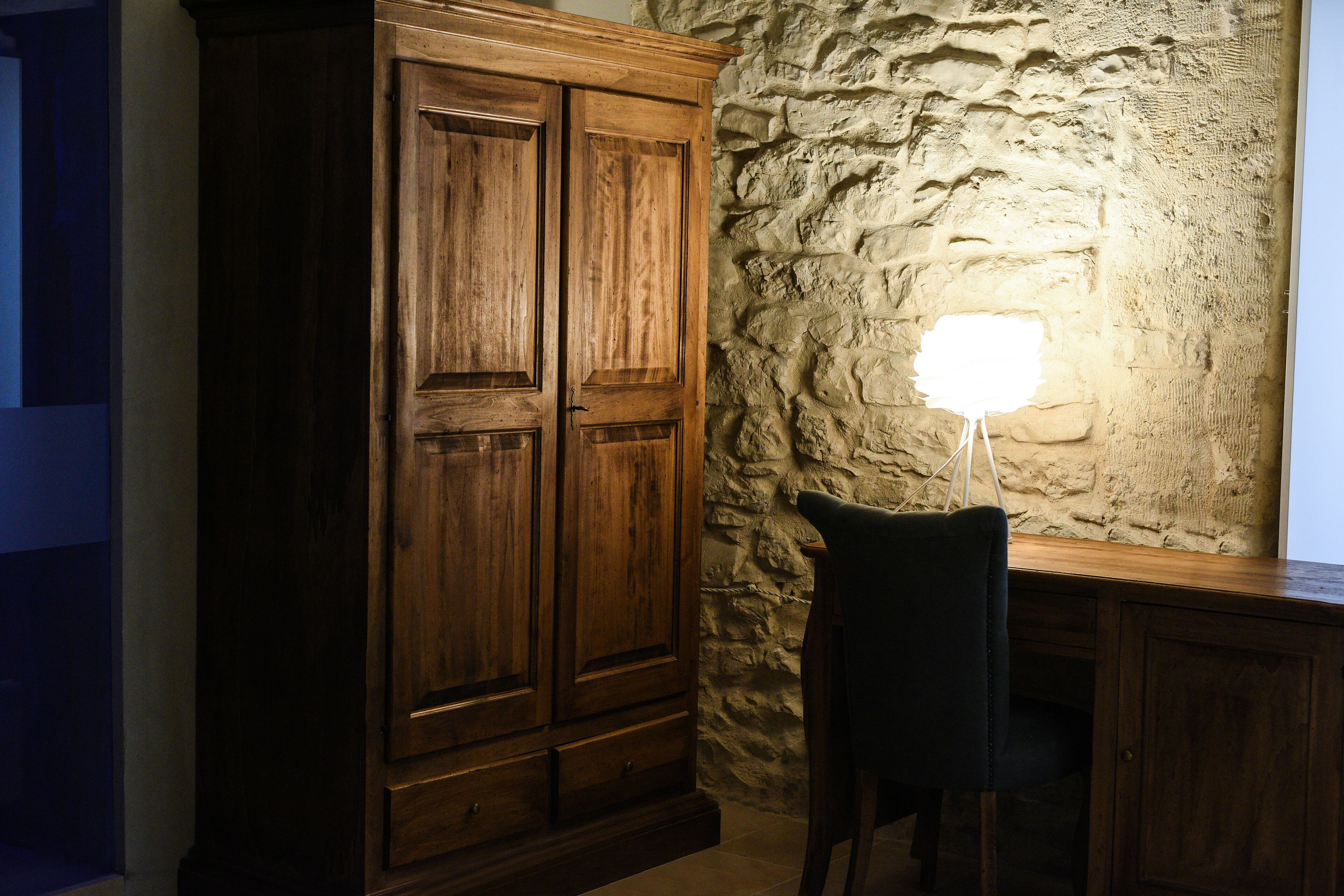 A room with a wooden wardrobe, a stone wall, and a desk with a chair.