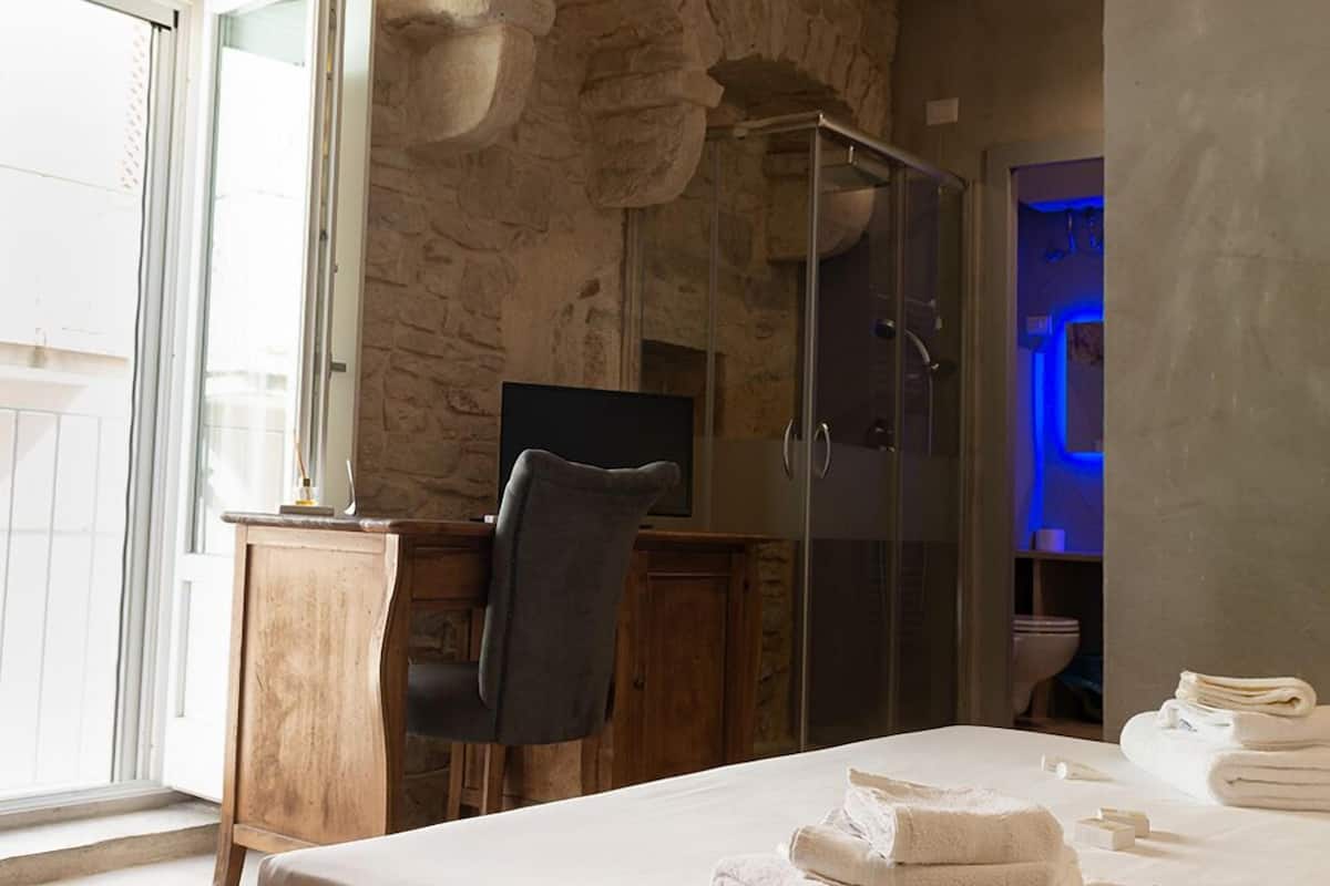 A bedroom with a stone wall, a wooden desk, a chair, a bed with towels, and a glass shower enclosure.