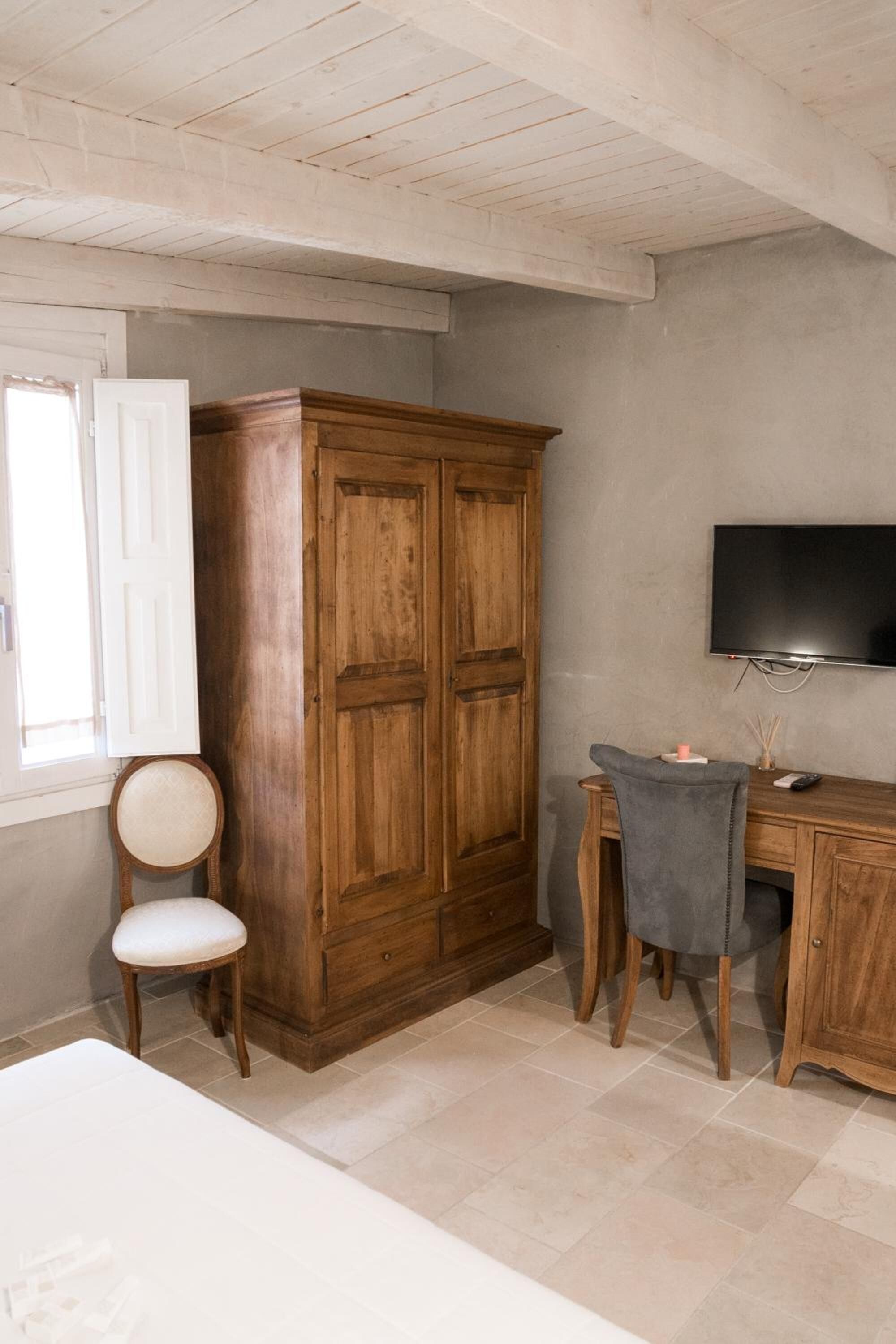 A room with a wooden wardrobe, a desk with a chair, a television, and a window with a curtain.