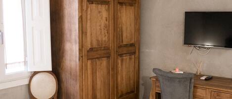 A room with a wooden wardrobe, a desk with a chair, a television, and a window with a curtain.