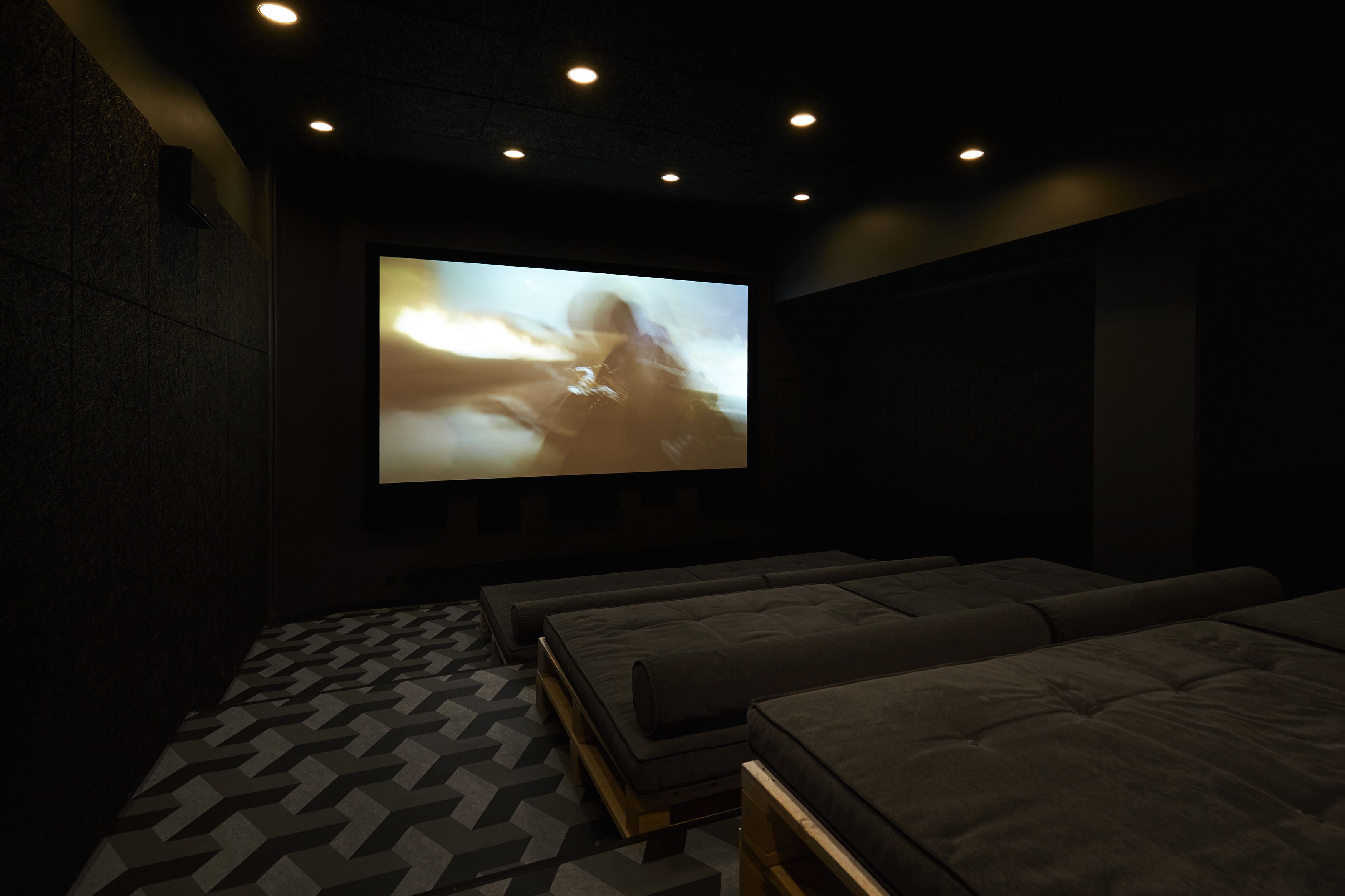 A dark room with a large screen displaying a blurred image, cushioned seating, and patterned flooring.