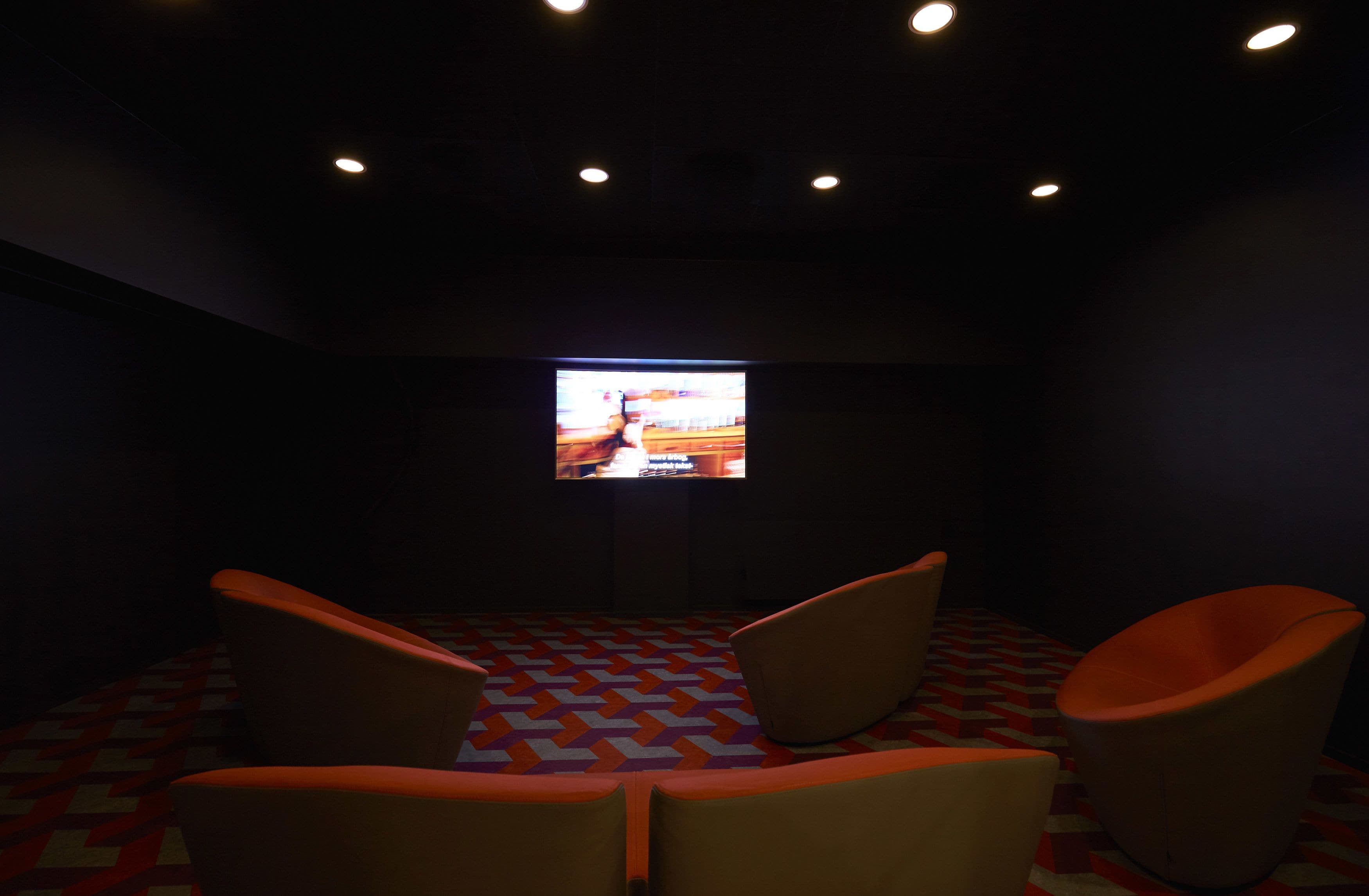 Une pièce sombre avec un grand écran diffusant une vidéo, deux fauteuils orange et un sol à motifs.