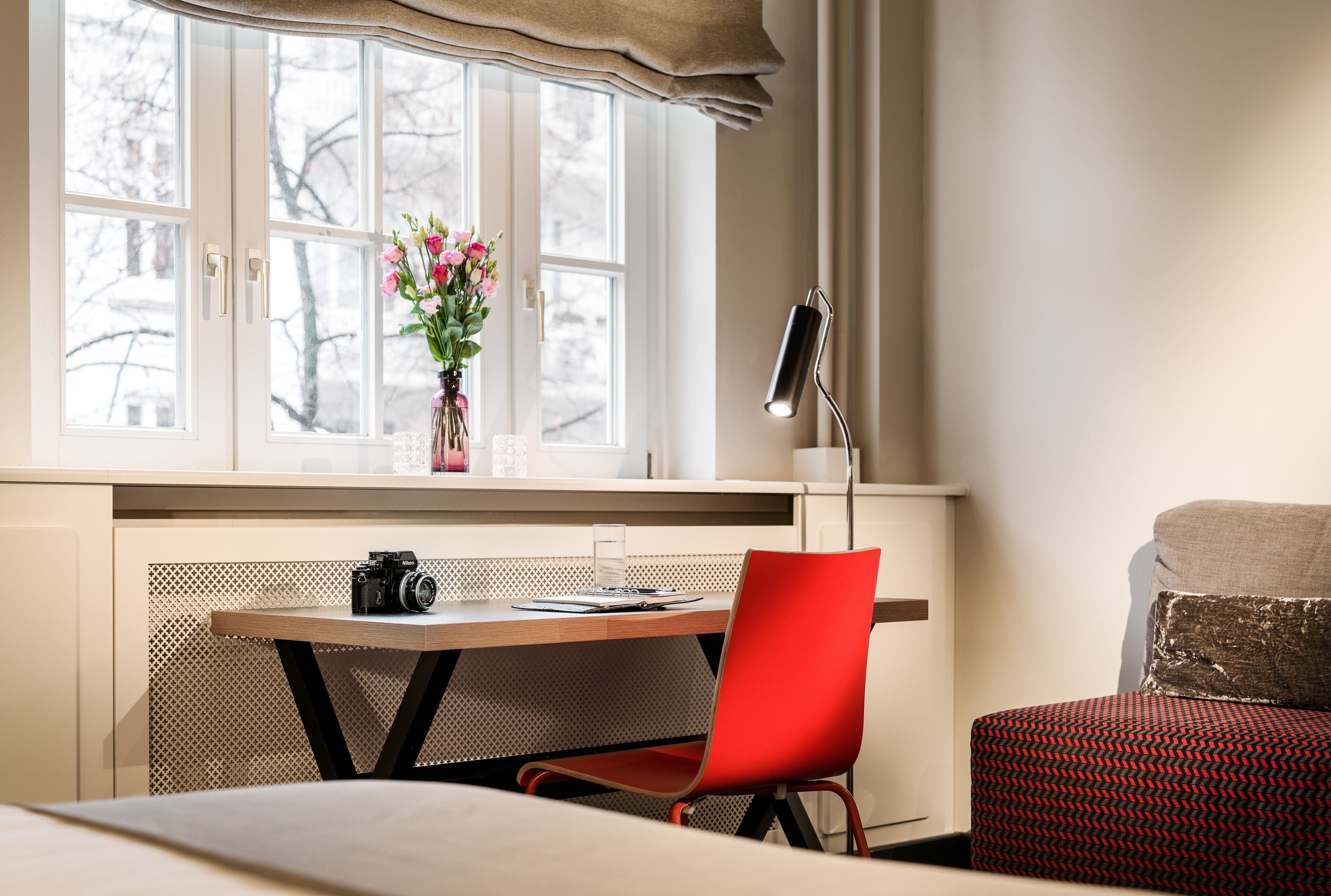 A room with a wooden desk, a red chair, a camera, a vase with flowers, and a lamp.