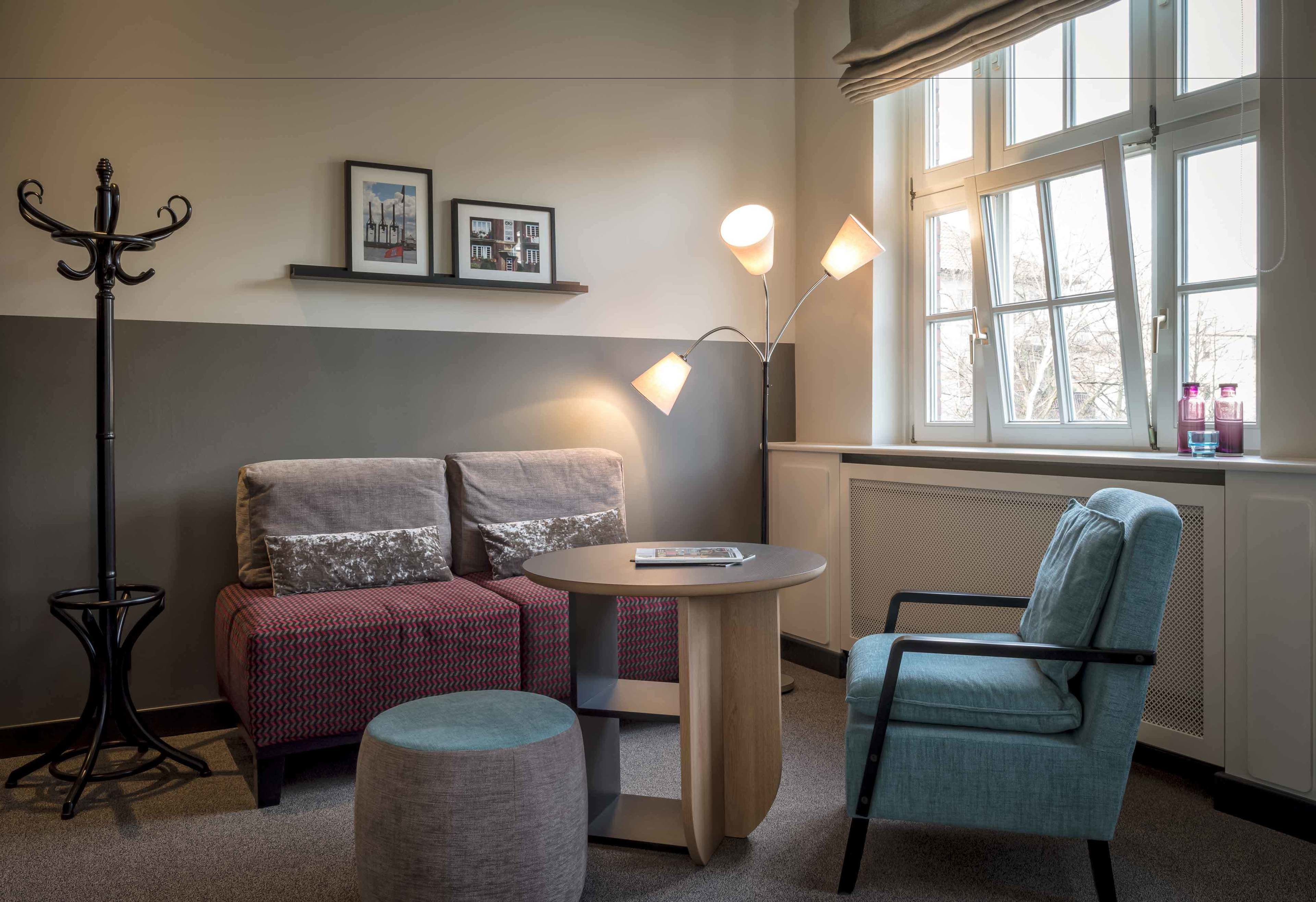 A cozy room with a sofa, a chair, a small table, and a floor lamp. There are two framed pictures on the wall and a window with curtains.