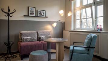 A cozy room with a sofa, a chair, a small table, and a floor lamp. There are two framed pictures on the wall and a window with curtains.