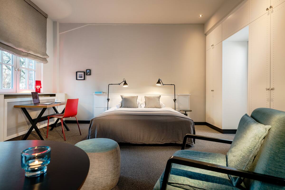 Habitación de hotel moderna con una cama, lámparas de noche, un escritorio con una computadora portátil, una silla roja y un reposapiés redondo.