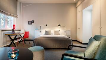 A modern hotel room with a bed, bedside lamps, a desk with a laptop, a red chair, and a round ottoman.
