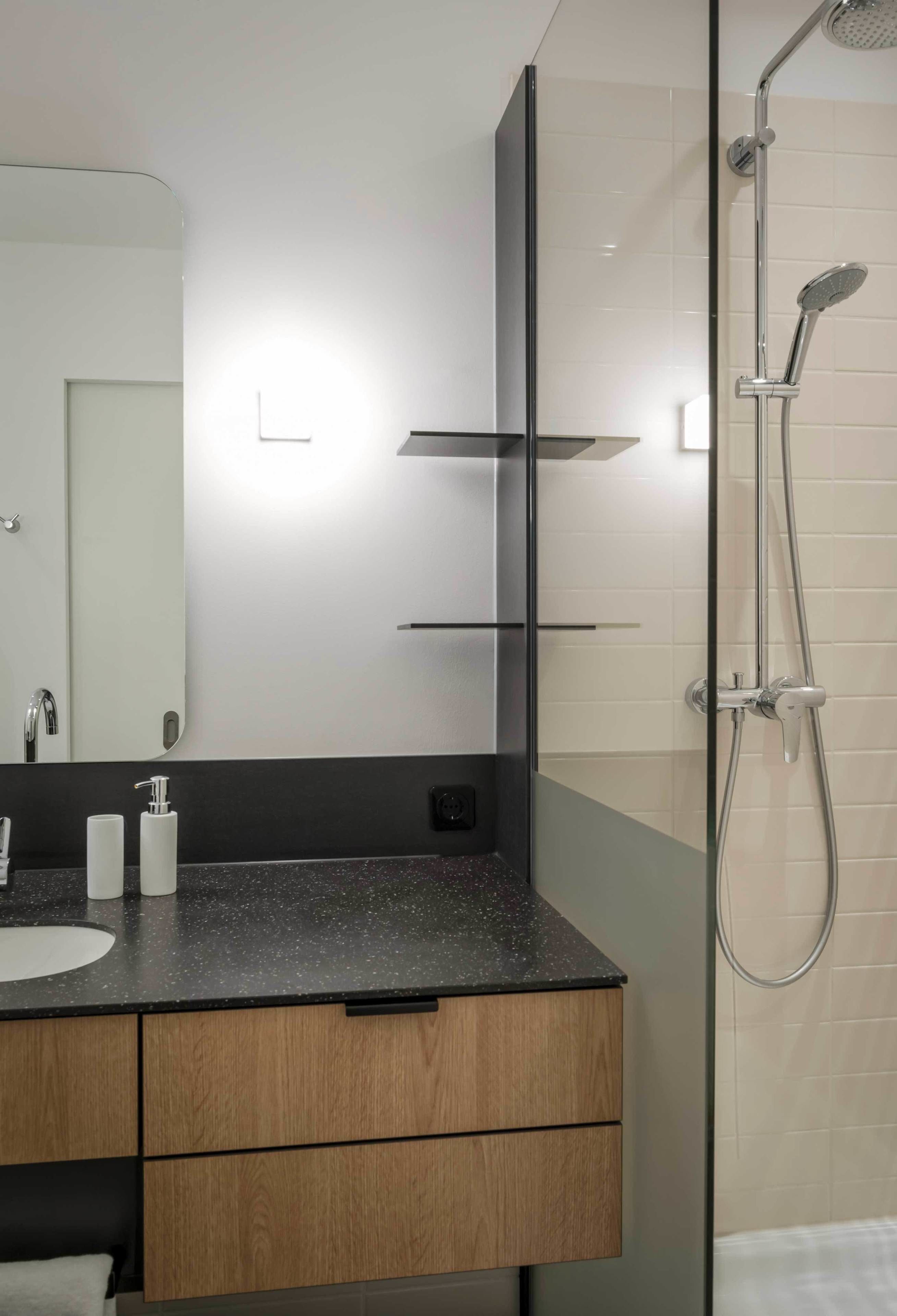 Une salle de bain moderne avec un grand miroir, un lavabo doté d’un plan de travail à motifs, des meubles en bois et une douche séparée par une paroi en verre.