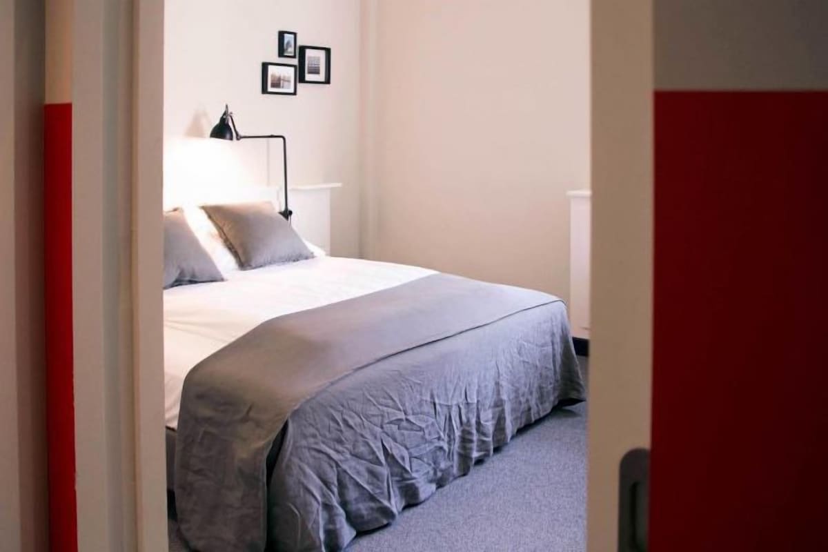 A neatly made bed with a grey duvet, a bedside lamp, and a wall with framed pictures.