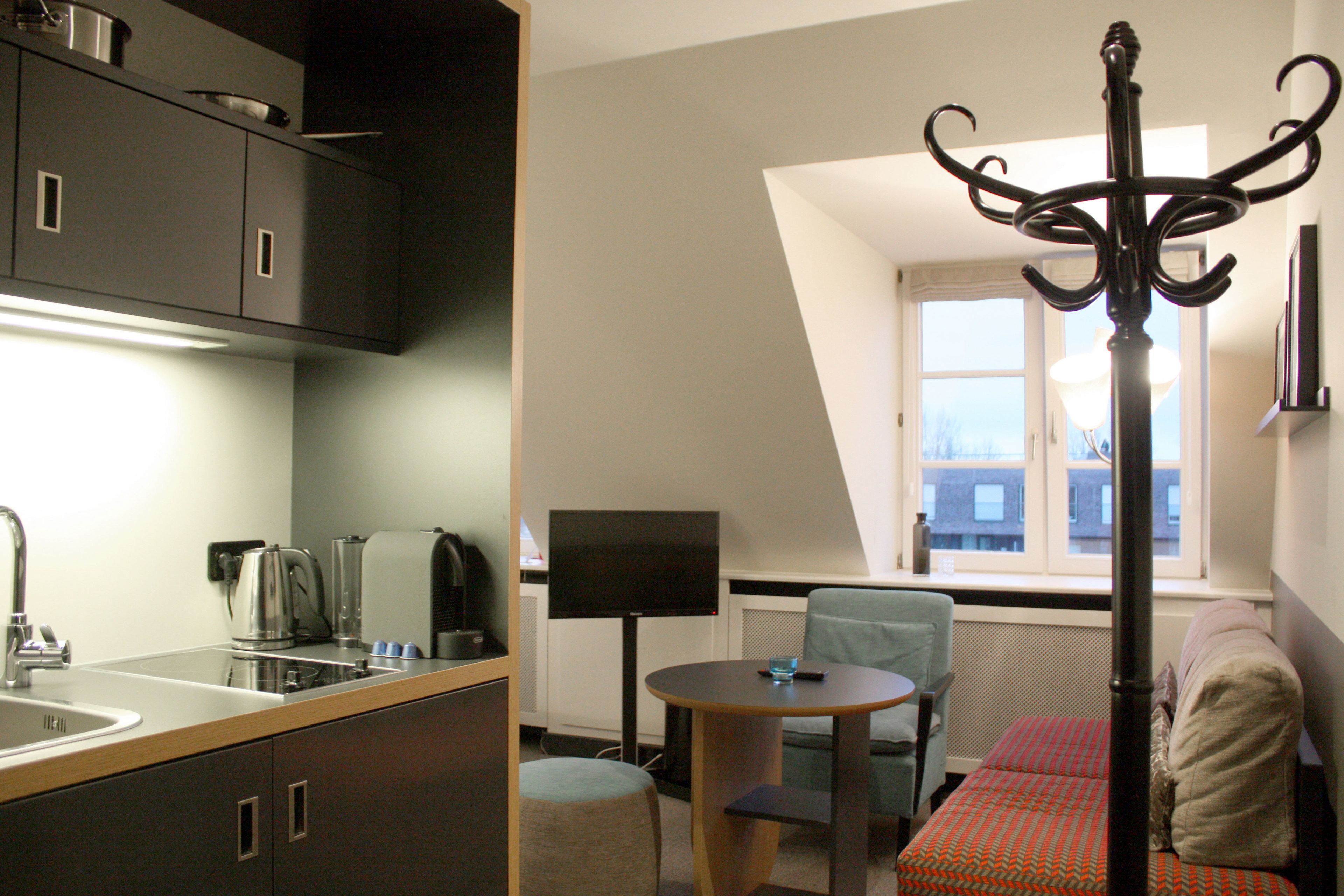 Quarto de hotel compacto com cozinha compacta, área de refeições com mesa e cadeiras, cama com colcha estampada e uma janela grande com vista para edifícios.