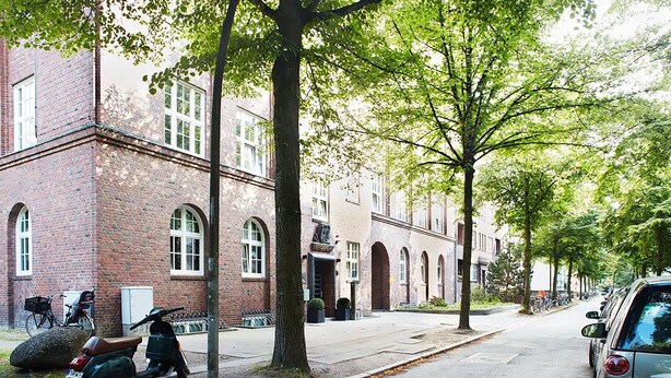 A street with a brick building, trees, parked scooters, and a car. - Apartment040 (Hamburg)