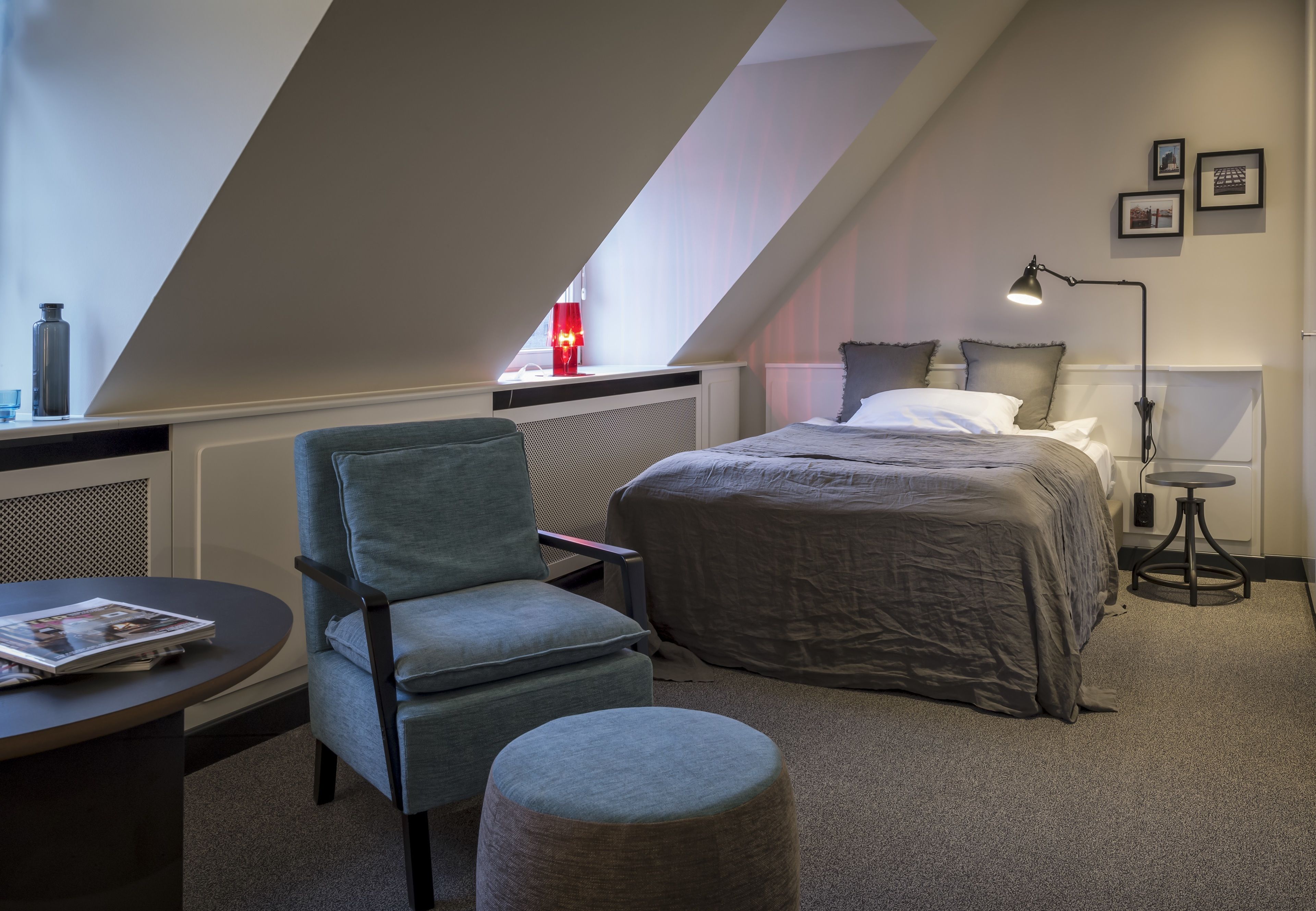 A modern bedroom with a sloped ceiling, a bed with grey bedding, a grey armchair, a small round ottoman, a bedside table with a lamp, and a radiator.
