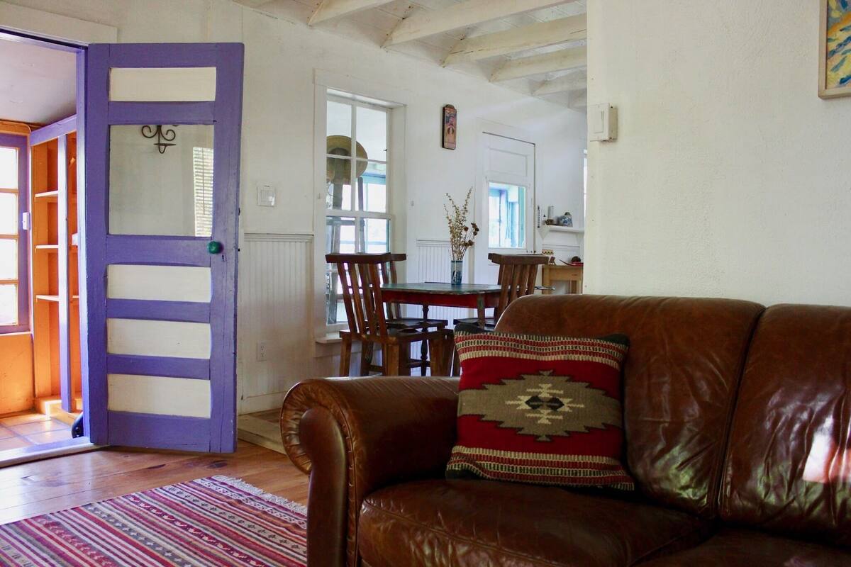 Una sala de estar con un sofá de cuero marrón, una puerta morada y un comedor con mobiliario de madera.