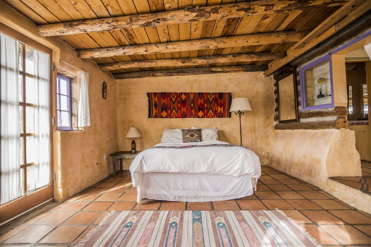 Un dormitorio con techo de madera, cama con ropa de cama blanca, tapiz colorido en la pared y suelo de baldosas.