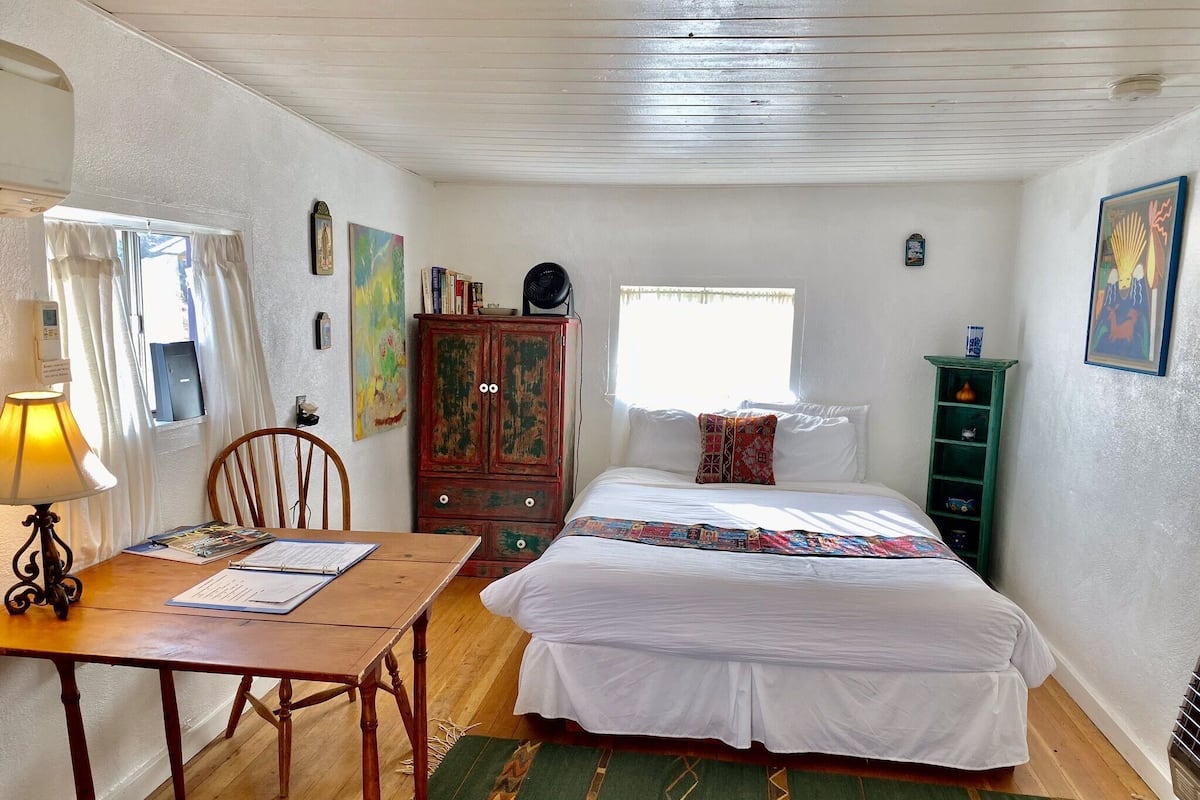 Un dormitorio con una cama de madera, un escritorio con una lámpara, una silla, un armario y una ventana con cortinas.