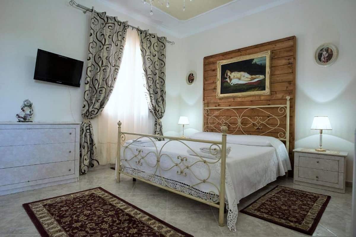 Une chambre à coucher avec un lit, des tables de chevet, une télévision, des rideaux, un tableau et un lustre.