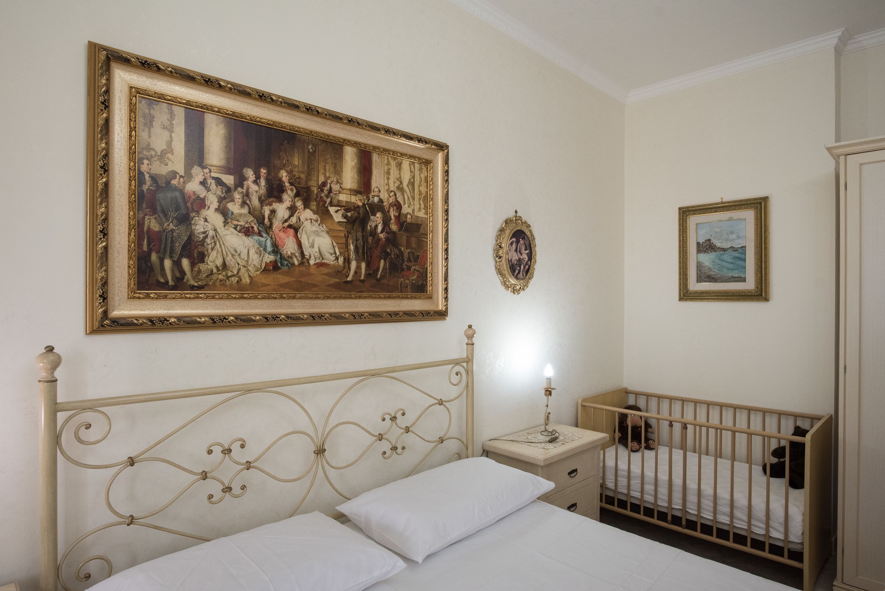 A bedroom with a large classical painting, a bed with white linens, a nightstand with a lamp, and a crib.
