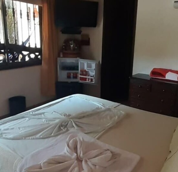Une chambre d’hôtel avec un lit, un téléviseur fixé au mur, une petite étagère avec quelques objets et une porte.