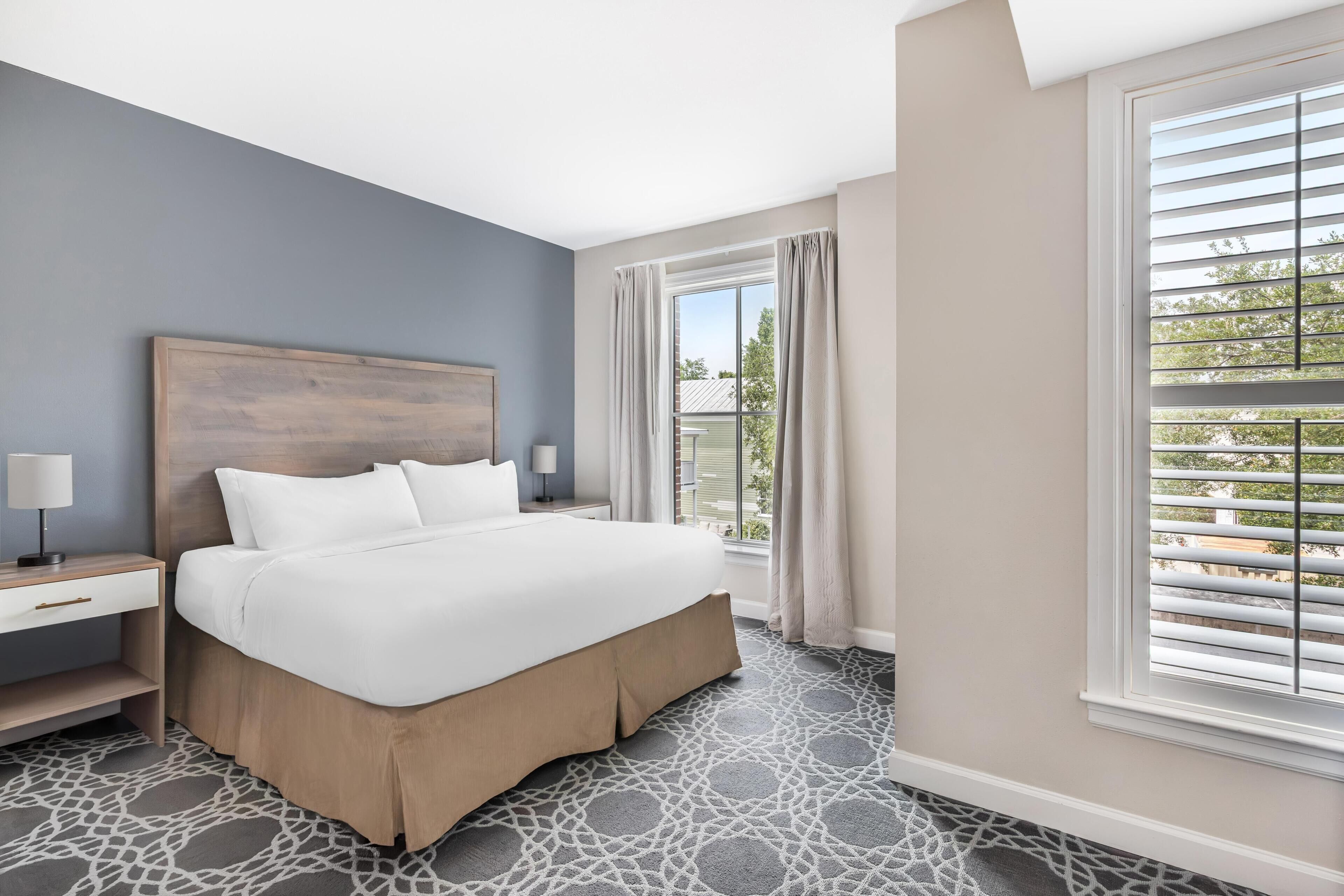 A hotel room with a large bed, a nightstand, a window with blinds, and a view of the outdoors.