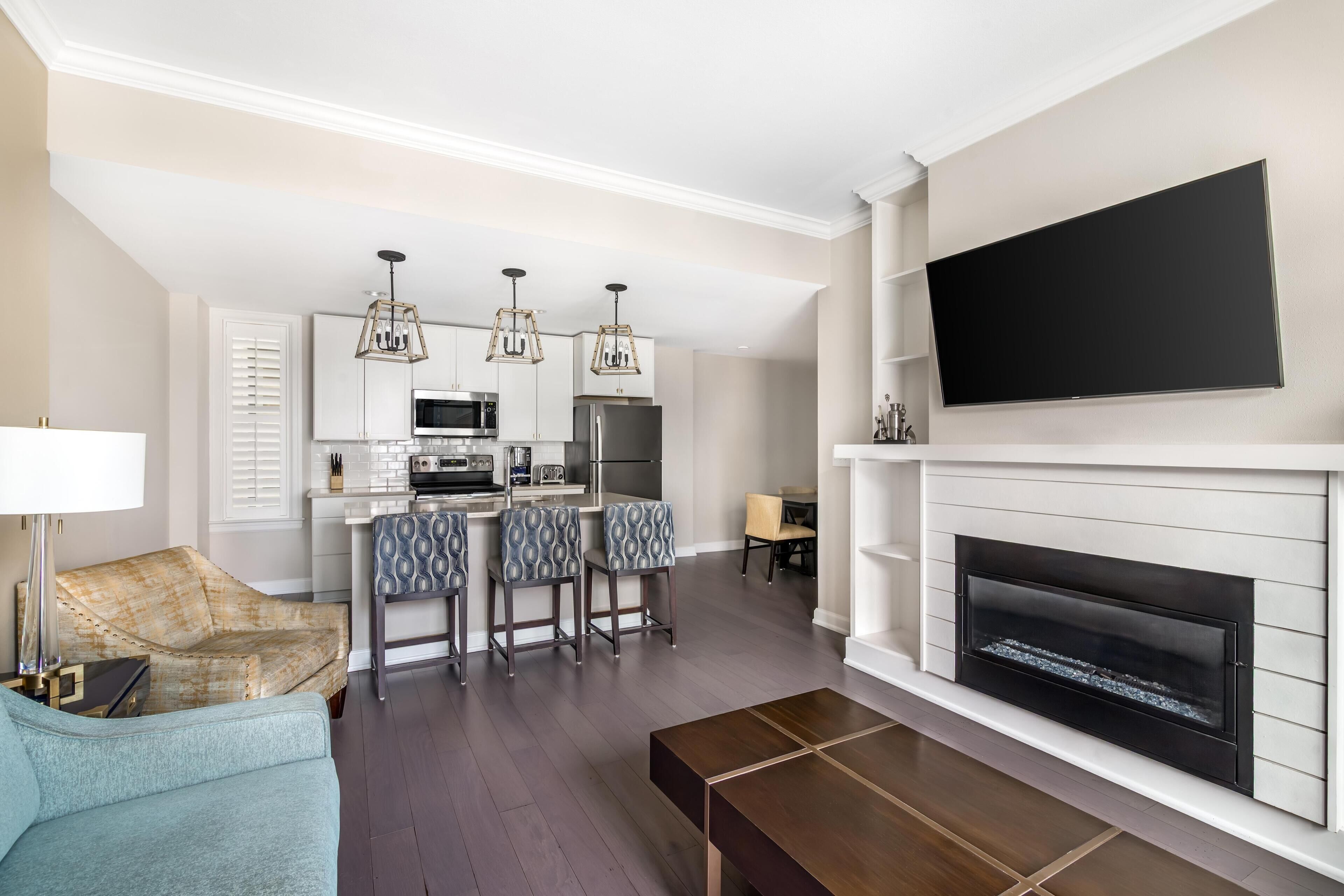 A modern living room with a fireplace, a flat-screen TV mounted above it, a kitchen area with white cabinetry and a central island, and a seating area with a sofa and a coffee table.