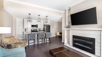 A modern living room with a fireplace, a flat-screen TV mounted above it, a kitchen area with white cabinetry and a central island, and a seating area with a sofa and a coffee table.