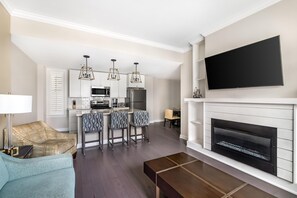 A modern living room with a fireplace, a flat-screen TV mounted above it, a kitchen area with white cabinetry and a central island, and a seating area with a sofa and a coffee table.