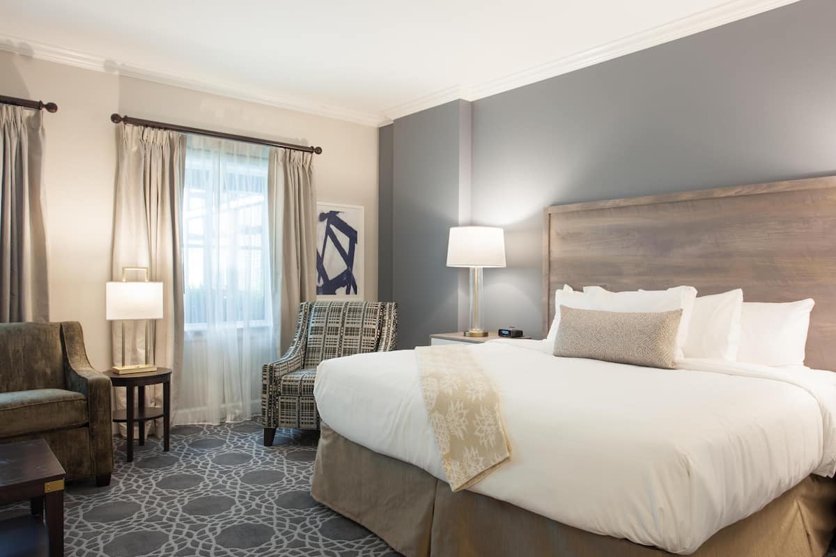 Habitación de hotel con una cama grande, una silla, una lámpara y vistas al exterior.
