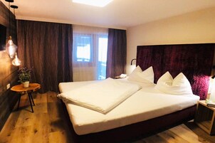 Habitación de hotel con una cama grande, piso de madera, una mesita con una planta y una ventana con cortinas.