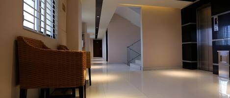 A modern interior with a hallway leading to a staircase, a seating area with a wicker chair, and a large window with blinds.