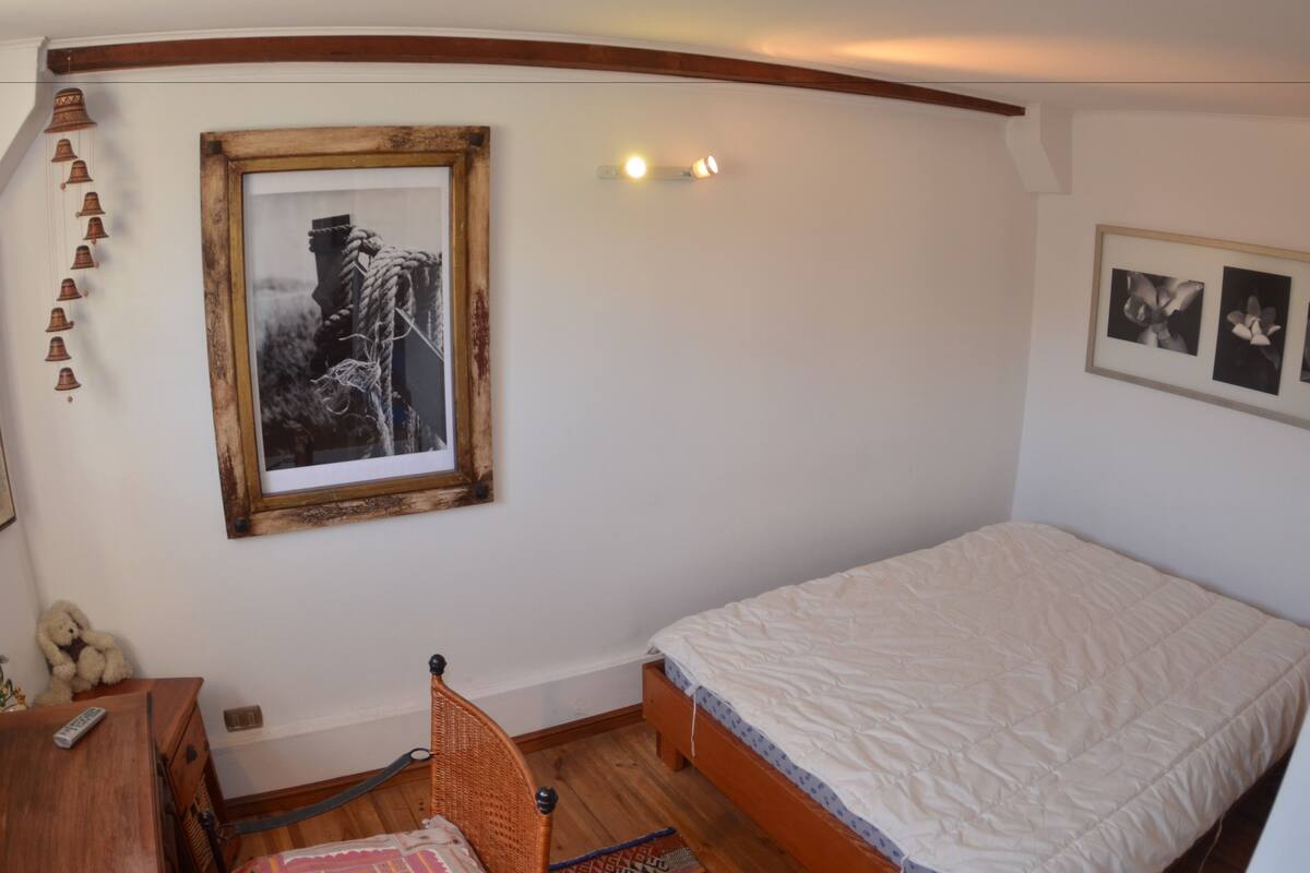 Ein Schlafzimmer mit einem Bett, einer Holzkommode, einem Sessel, einem gerahmten Bild und einer Wandleuchte.