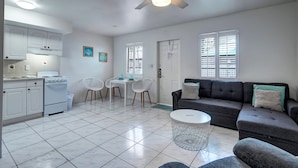Une cuisine moderne avec des armoires blanches, une cuisinière et un ventilateur de plafond. On y trouve un coin repas avec une table et des chaises, ainsi qu’un salon avec un canapé et une table basse.