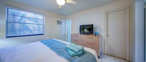 Un dormitorio con cama, ventilador de techo, televisor sobre una cómoda y ventana con persianas.