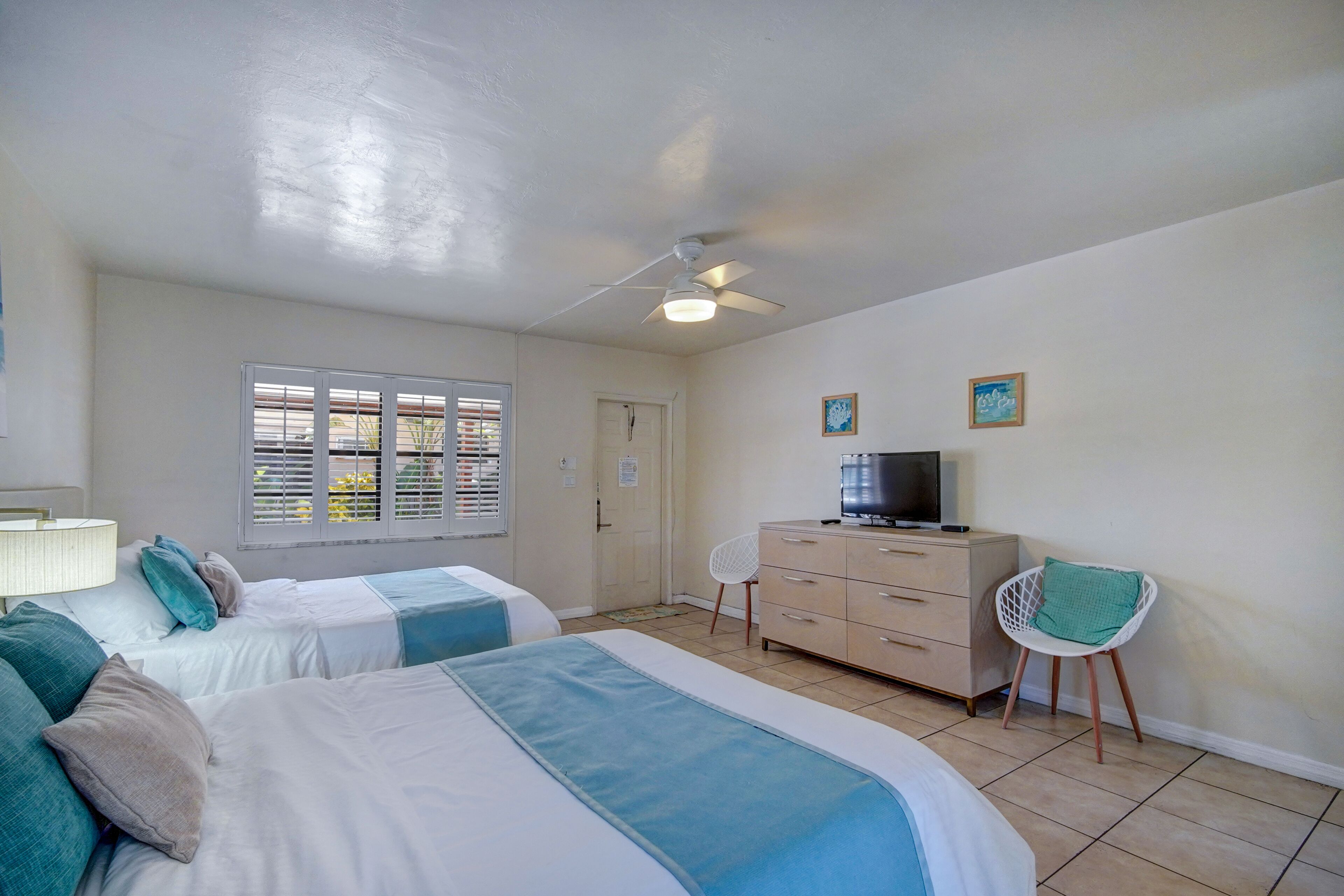 A bedroom with two beds, a ceiling fan, a TV on a dresser, and a window with blinds.