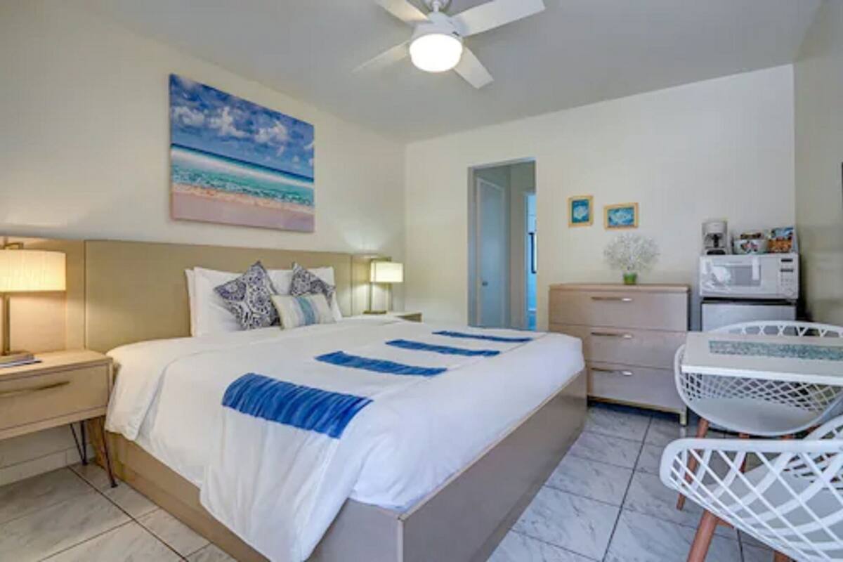 A hotel room with a bed, bedside tables, a desk, a chair, a ceiling fan, and a painting of a beach.