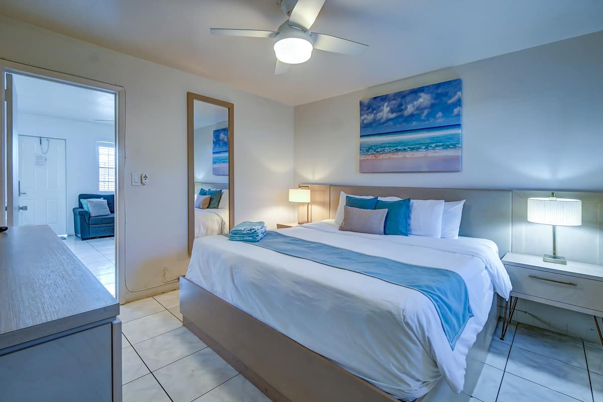 A hotel room with a bed, bedside tables, a ceiling fan, a painting of a beach, and a view of the adjacent room.