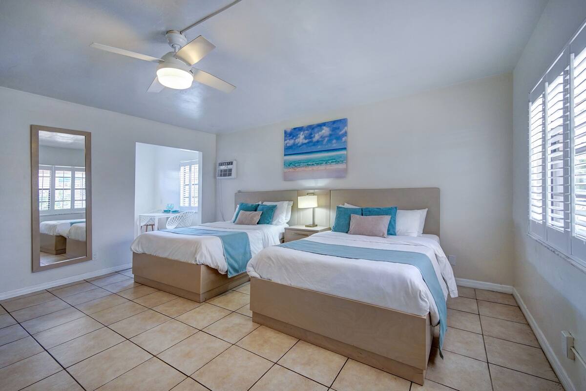 A hotel room with two beds, a ceiling fan, a beach-themed painting, and a window with blinds.