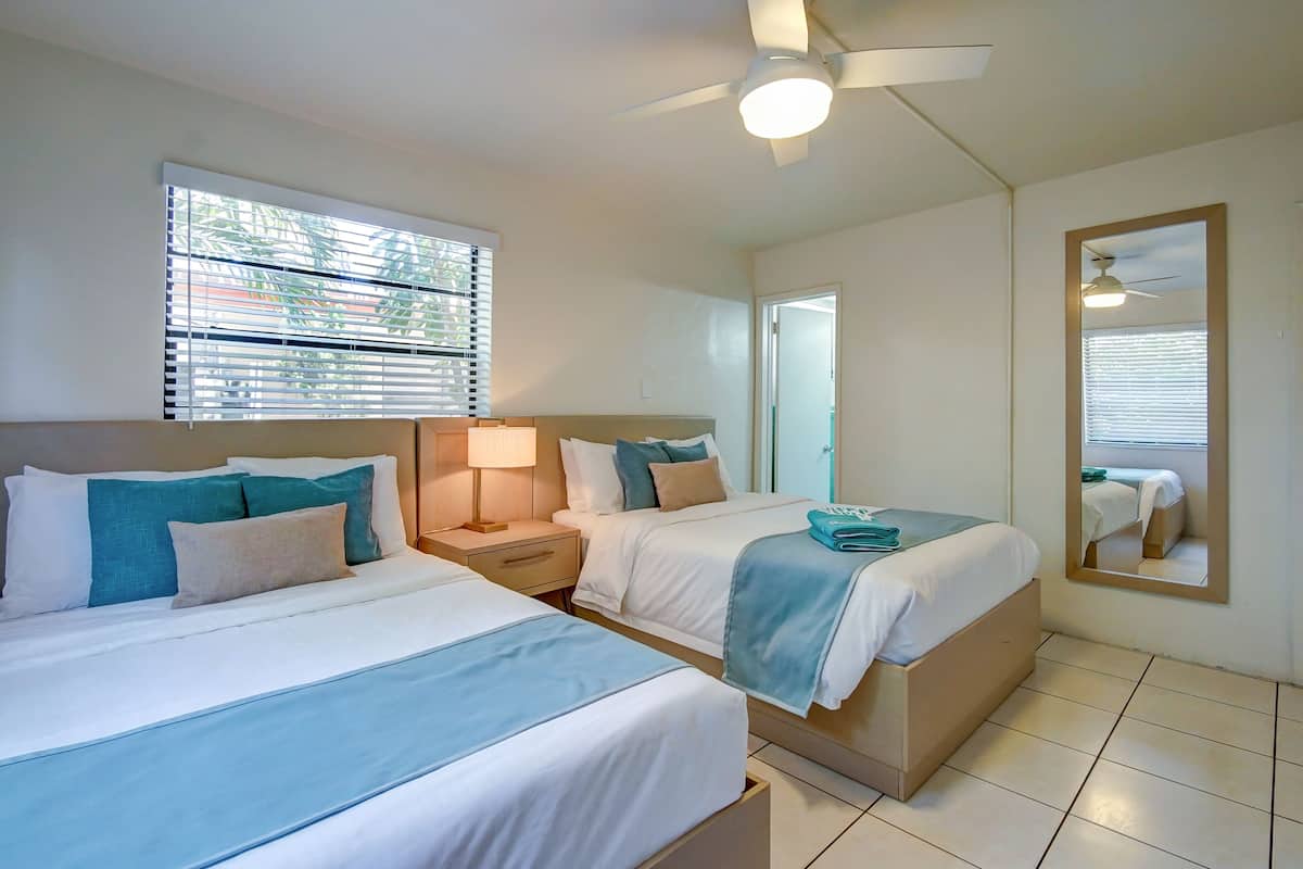 A hotel room with two beds, a ceiling fan, a window with blinds, a bedside table with a lamp, and a mirror on the wall.