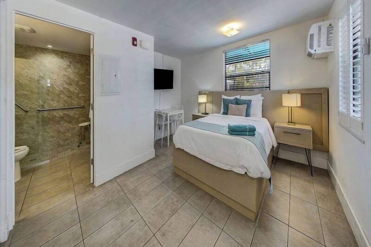 A hotel room with a bed, bedside table, TV, and a shower area.